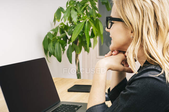 Die schöne blonde Kaukasierin arbeitet von ihrem Wohnzimmer aus mit ihrem Laptop auf einem Holztisch. Sie trägt schwarze Freizeitkleidung. — Stockfoto