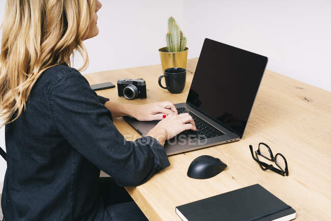 Hermosa mujer rubia caucásica trabaja desde su sala de estar con su computadora portátil en un escritorio de madera. Ella usa ropa casual negra. - foto de stock