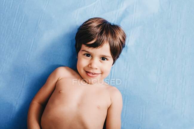 From above of cheerful shirtless little boy looking at camera while lying on blue hospital bed — Stock Photo
