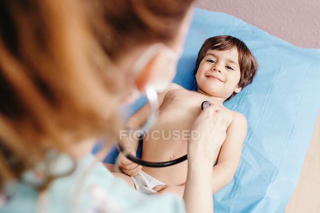 From above of cheerful little boy lying on couch and being examined by unrecognizable female doctor with stethoscope in modern clinic — Stock Photo