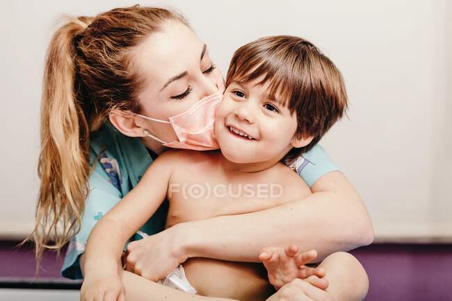 Cheerful young female in protective medical mask hugging and kissing cute laughing little patient — Stock Photo