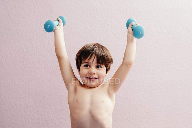Счастливый здоровый ребенок с голубыми гантелями в поднятых руках, стоящих у розовой стены и глядя в сторону — стоковое фото