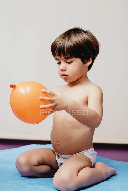 Triste niño sin camisa sentado en la cama del hospital y sosteniendo globo naranja, mientras que representa el concepto de enfermedad respiratoria y tratamiento - foto de stock