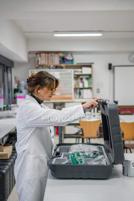 Вид сбоку на взрослую женщину в белом халате, открывающую кейс с новым оборудованием во время работы в современной лаборатории — стоковое фото