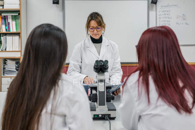 Donna adulta in vestaglia bianca che legge appunti vicino al microscopio mentre insegna alle giovani donne nel moderno laboratorio dell'università — Foto stock