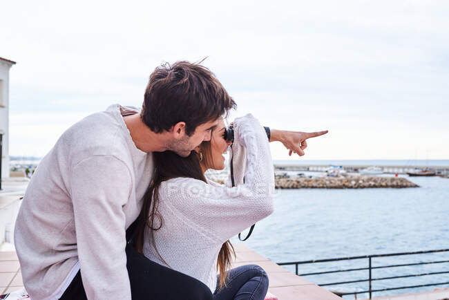 Vista laterale di allegra coppia romantica in abbigliamento casual seduta sulla terrazza con binocolo e godendo della vista mentre trascorrete le vacanze insieme al mare — Foto stock