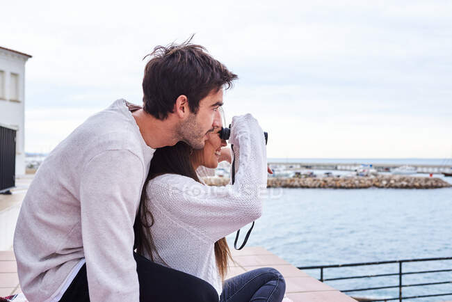 Vista lateral de la alegre pareja romántica en ropa casual sentada en la terraza con prismáticos y disfrutando de la vista mientras pasan las vacaciones juntos en la playa - foto de stock