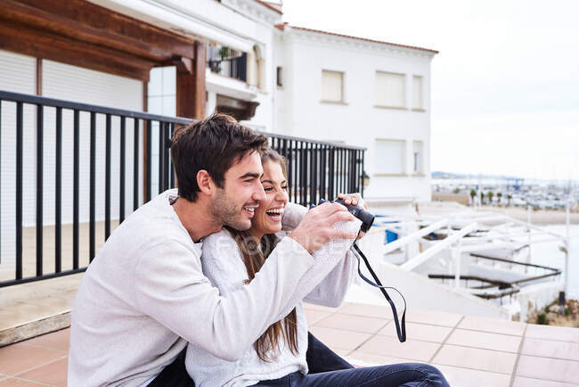 Vue latérale d'un couple romantique joyeux en tenue décontractée assis sur la terrasse avec jumelles et bénéficiant d'une vue tout en passant des vacances ensemble au bord de la mer — Photo de stock