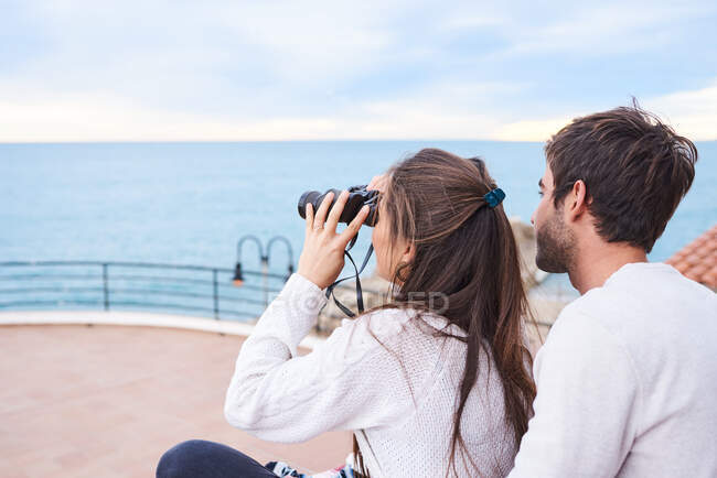 Seitenansicht eines fröhlichen romantischen Paares in Freizeitkleidung, das mit Fernglas auf der Terrasse sitzt und die Aussicht genießt, während es gemeinsam Urlaub am Meer verbringt — Stockfoto