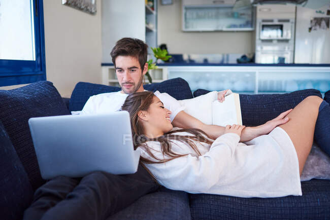 Alto ángulo de hombre joven navegando portátil y mujer leyendo libro interesante mientras descansan juntos en cómodo sofá en la sala de estar moderna - foto de stock