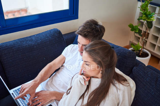 Високий кут перегляду молодого чоловіка ноутбук і жінка читають цікаву книгу, відпочиваючи разом на зручному дивані в сучасній вітальні — стокове фото