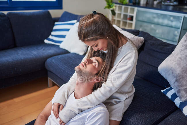 Mujer joven positiva abrazando al hombre feliz mientras está sentado en el sofá en la acogedora sala de estar - foto de stock