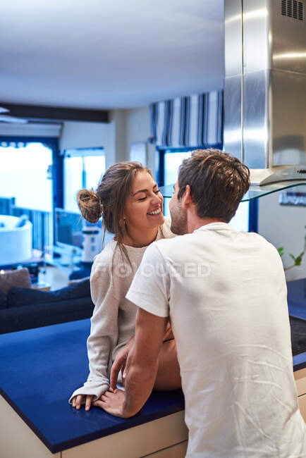 Vista lateral de la joven feliz sentada en el mostrador de la cocina y abrazando al esposo cariñoso mientras pasan el día juntos en un apartamento moderno - foto de stock