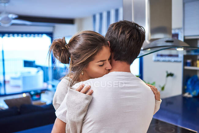 Side view of happy young woman sitting on kitchen counter and embracing loving husband while spending day together in modern apartment — Stock Photo