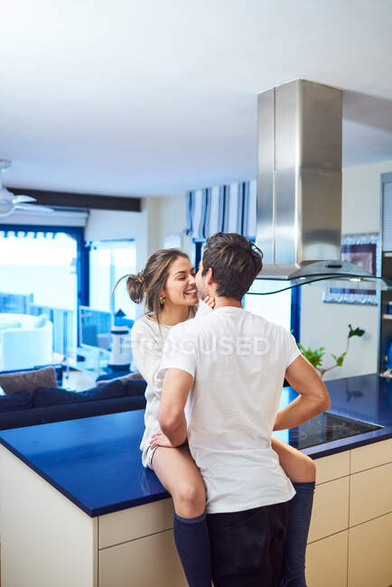 Seitenansicht einer glücklichen jungen Frau, die auf dem Küchentisch sitzt und ihren liebevollen Mann umarmt, während sie den Tag zusammen in einer modernen Wohnung verbringt — Stockfoto