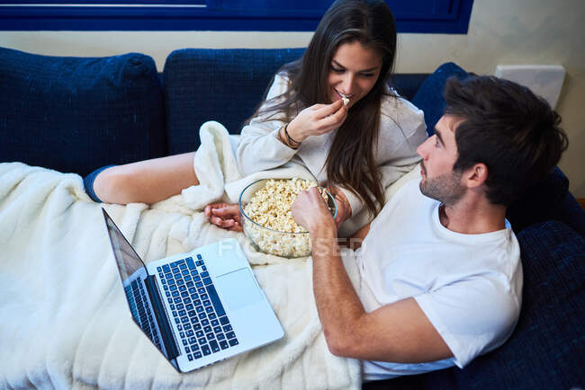 Desde arriba de alegre joven hombre y mujer en ropa casual comer palomitas de maíz y ver la película en el ordenador portátil mientras descansan juntos en el sofá acogedor en casa - foto de stock