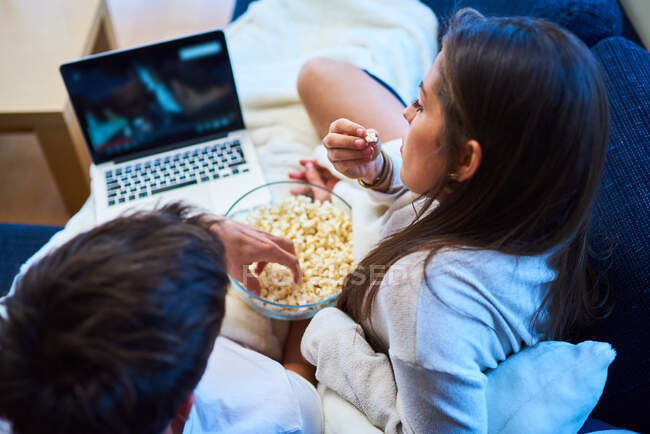Веселый молодой человек и женщина в повседневной одежде едят попкорн и смотрят фильм на ноутбуке, отдыхая вместе на уютном диване дома — стоковое фото