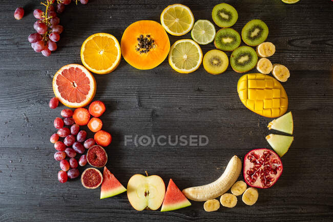 Vista superior de varias frutas y verduras sanas peladas y cortadas dispuestas en círculo sobre una mesa de madera negra - foto de stock