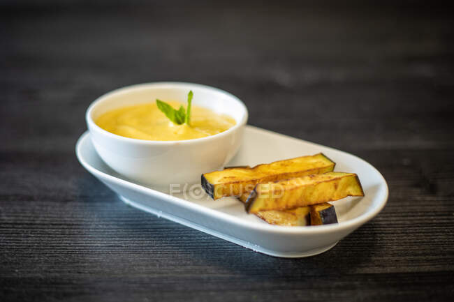 Bowl of delicious puree with herb placed on ceramic plate near pieces of fried eggplant on black wooden table in cafe — Stock Photo