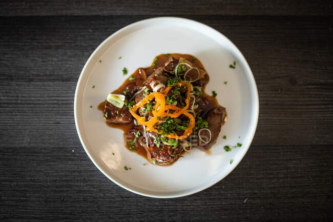 Верхний вид восхитительного жареного мяса подается с соусом и травами на керамической тарелке на черном деревянном столе — стоковое фото