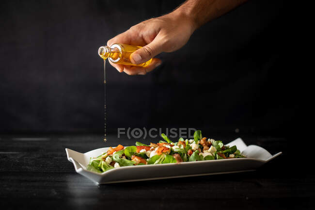 Chef irreconocible derramando aceite de una pequeña botella sobre ensalada de espinacas sanas con tomates y champiñones sobre fondo negro - foto de stock