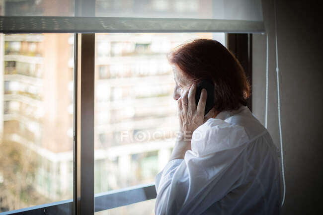 Side view of senior female in white blouse having phone call while standing near window in hospital ward visiting relative — Stock Photo