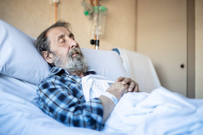 Спокійний літній чоловік з бородою лежить під ковдрою на ліжку в лікарняній палаті і спить — стокове фото