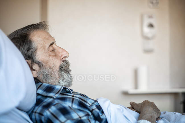 Спокійний літній чоловік з бородою лежить під ковдрою на ліжку у лікарні й спить. — стокове фото