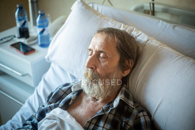 Hombre de edad calma con barba acostado debajo de la manta en la cama en la sala de hospital y durmiendo - foto de stock