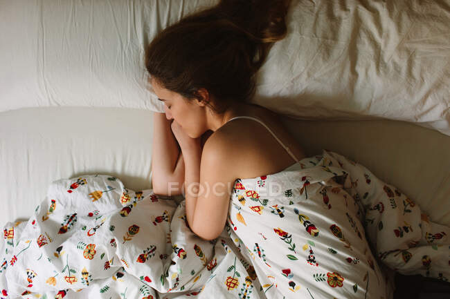 Desde arriba vista lateral de la joven hembra serena con el pelo ondulado largo con sujetador de encaje durmiendo en la cama acogedora con sábanas blancas y manta ornamental - foto de stock