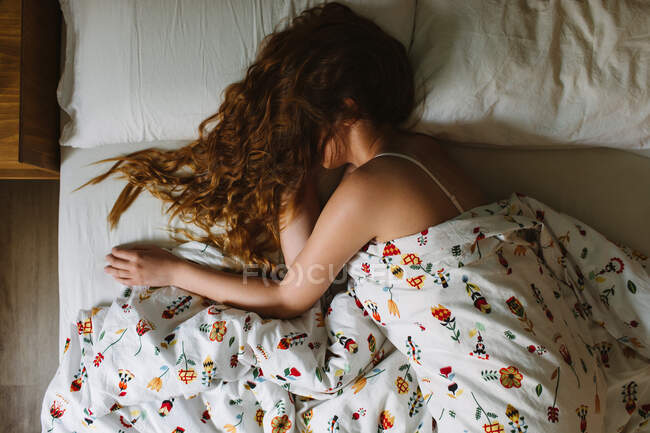 De cima vista lateral da fêmea serena irreconhecível com cabelos longos ondulados sobre o rosto usando sutiã de renda dormindo em cama acolhedora com lençóis brancos e cobertor ornamental — Fotografia de Stock