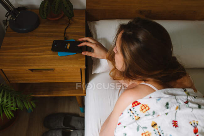 From above top view of young female lying in bed and turning off alarm on mobile phone after awakening in cozy bedroom — Stock Photo