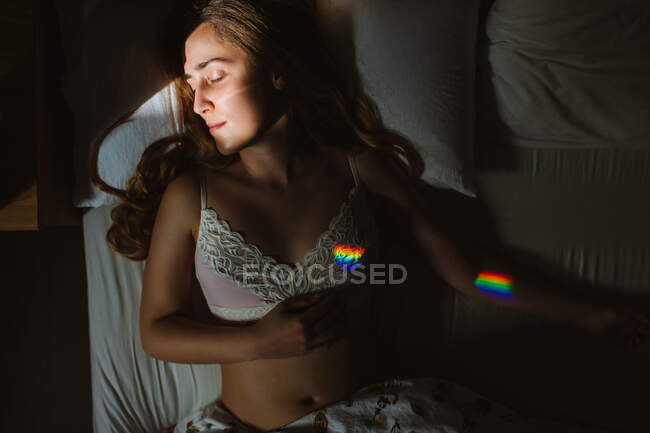 Von oben eine junge sinnliche Frau in weißem SpitzenbH mit buntem Regenbogen mit geschlossenen Augen, die morgens im Bett schläft — Stockfoto