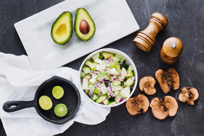 Top view of bowl with delicious avocado salad in composition with lime halves in skillet and cut avocado with fried apple chips on table — Stock Photo