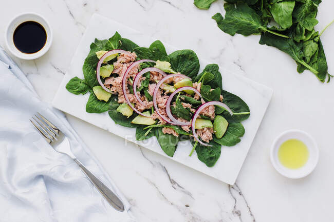 Overhead view of flat plate with chopped avocado and tuna with red onion on fresh green spinach on white marble table — Stock Photo