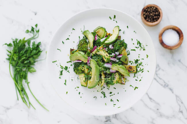 From above of round white plate with served pile of avocado salad with zucchini and broccoli garnished with chopped red onion and parsley — Stock Photo