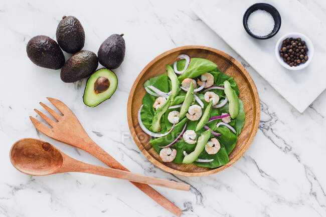 Flat lay of round wooden plate with salad of green and avocado garnished with red onion and shrimps composed with ingredients on white marble — Stock Photo