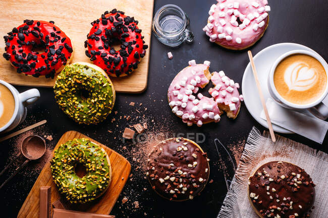 Flat lay of various doughnuts with sweet toppings and chocolate bars composed with cup of cappuccino on black table — Stock Photo