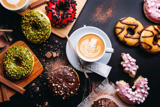 Плоская кладка различных пончиков со сладкими начинками и шоколадные батончики, составленные с чашкой капучино на черном столе — стоковое фото