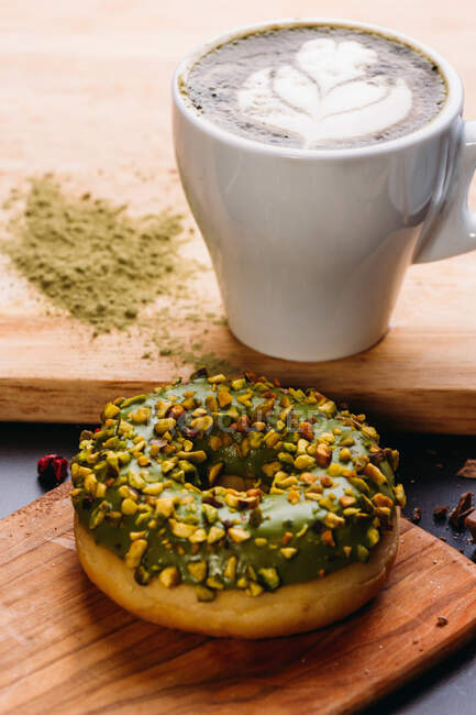 High angle of delicious doughnut with green topping and chopped nuts placed on wooden board with cup of aromatic cappuccino — Stock Photo