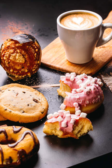 Різні пончики з солодкими начинками та шоколадними батончиками, складені з чашкою капучино на чорному столі — стокове фото