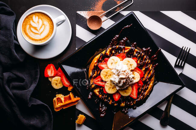 Вид на капучино в белой кружке на столе с тарелкой круглых вафель с бананом и клубникой, увенчанной шоколадным соусом и взбитыми сливками — стоковое фото