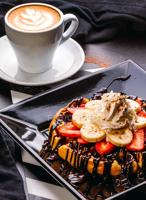 From above cappuccino in white mug on table with plate of round waffle with banana and strawberry topped with chocolate sauce and whipped cream — Stock Photo