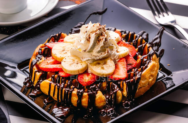 Сверху тарелка с круглой вафлей с бананом и клубникой, увенчанной шоколадным соусом и взбитыми сливками — стоковое фото