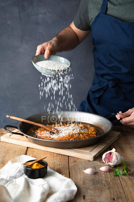 Crop man en tablier bleu foncé ajoutant du riz blanc aux ingrédients rôtis hachés sur une grande poêle en métal tout en préparant un délicieux plat salé à la maison — Photo de stock
