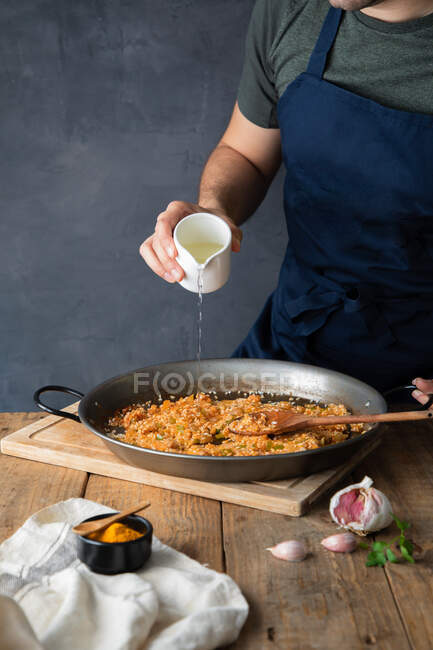 Homem de colheita em avental azul escuro derramando óleo de arroz com ingredientes torrados picados em grande panela de metal enquanto cozinha prato salgado apetitoso em casa — Fotografia de Stock