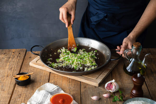 High angle of crop cook in dark apron stirring with wooden spatula mix of chopped green chili pepper and onion on big metal pan while adding oil before firing in contemporary kitchen at home — Stock Photo