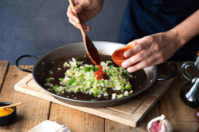Von oben nicht wiederzuerkennende Köchin fügt Tomatenmark mit Holzspatel zu gehackter grüner Chilischote und Zwiebeln hinzu, die auf einer großen Metallpfanne gemischt werden, während sie sich darauf vorbereitet, Zutaten in der modernen Küche zu Hause zu braten. — Stockfoto