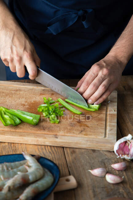 Desde arriba, el chef corta ají verde en una tabla de cortar de madera mientras prepara condimentos de especias para un delicioso plato con la adición de ajo y camarones crudos frescos en la mesa rústica - foto de stock