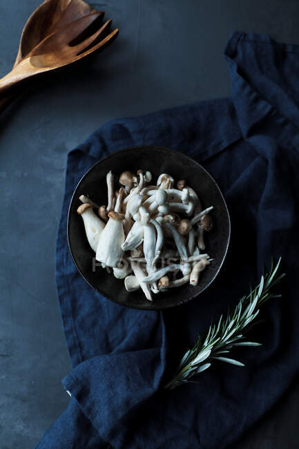 Vista superior da tigela com cogumelos não cozidos e alecrim verde fresco decorre na toalha azul escuro em arranjo com colher de madeira e garfo na mesa preta na cozinha rústica — Fotografia de Stock
